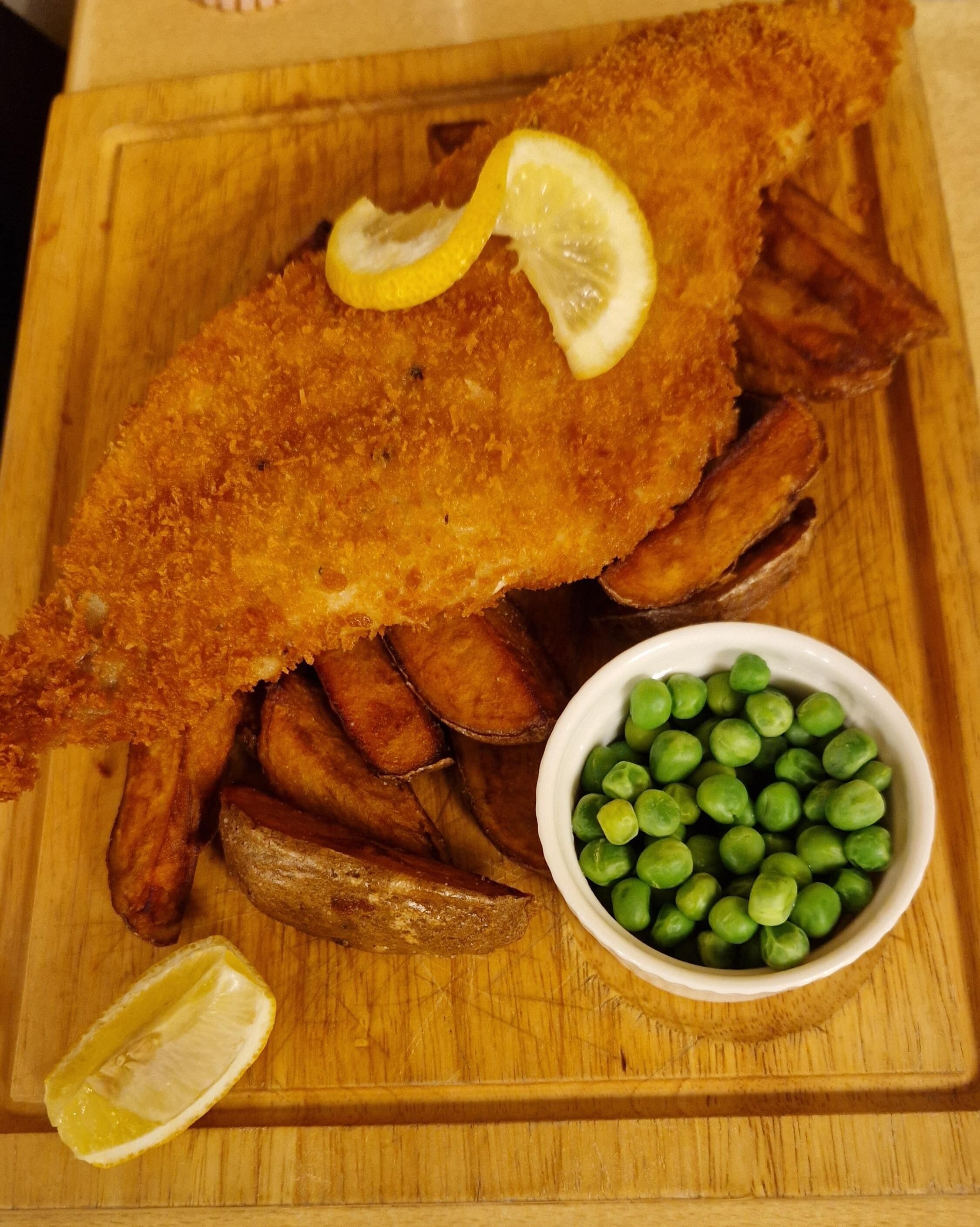 Un plato con un filete de pescado empanizado, patatas asadas y guisantes verdes sobre una tabla de madera.