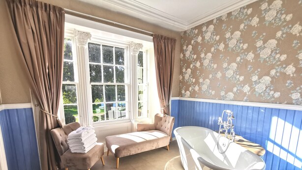A bathroom with a bathtub, two chairs, a window with curtains, and floral wallpaper. - Lunan House (Arbroath)