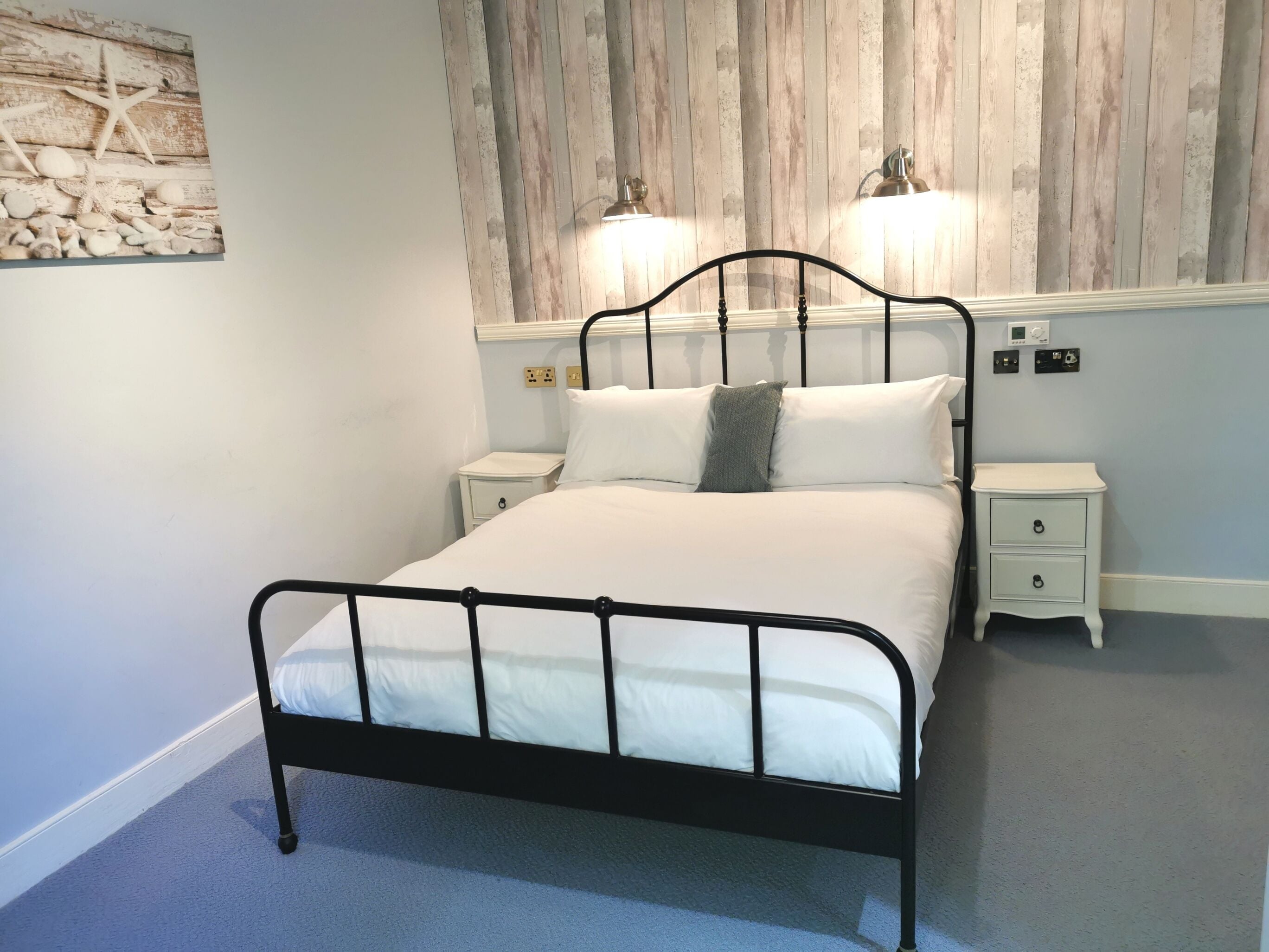 A bedroom with a black metal bed frame, white bedding, two bedside lamps, a small bedside table, and a wall-mounted artwork.