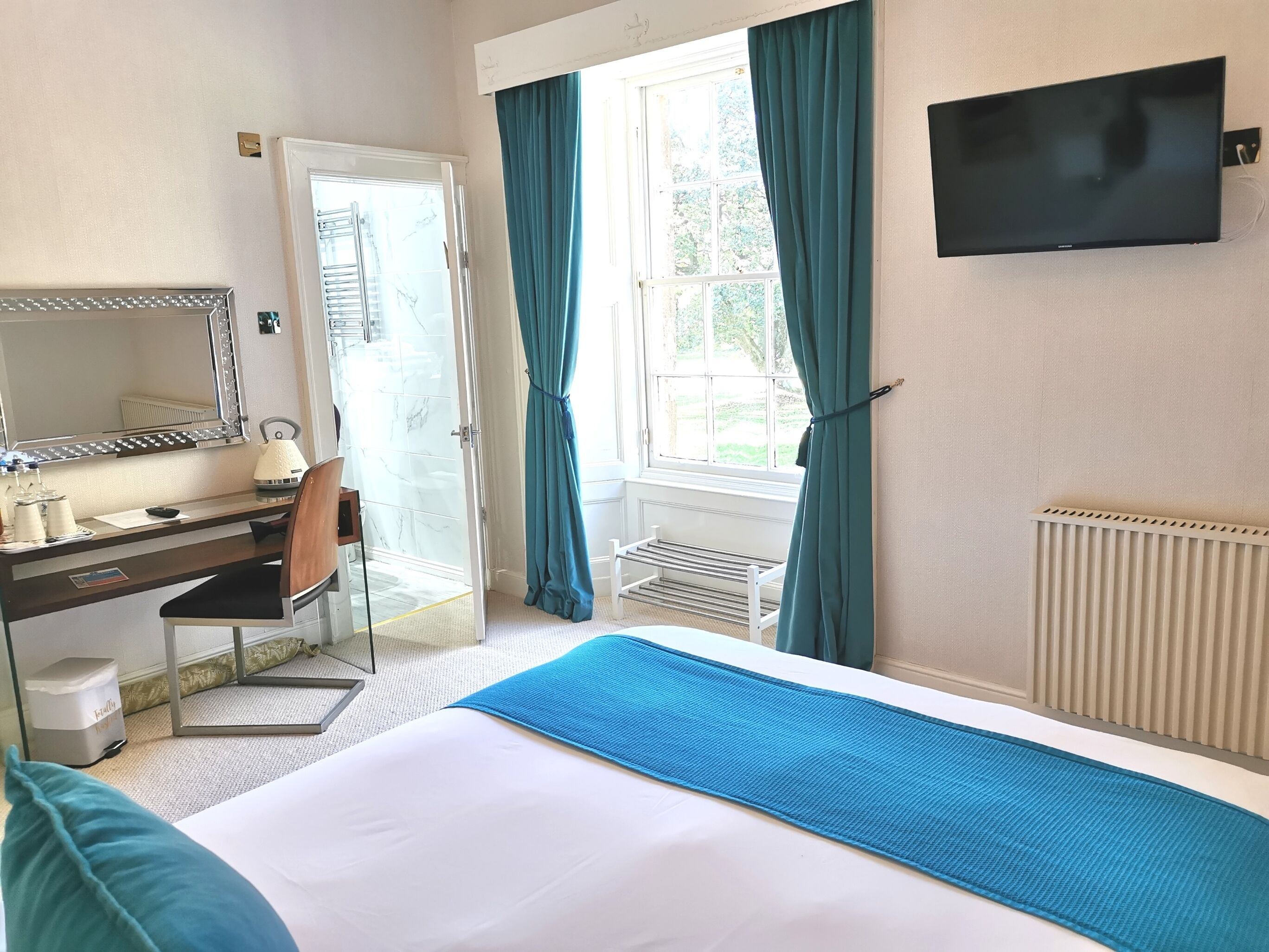 A hotel room with a bed, a desk with a chair, a mirror, a TV, and a window with curtains.