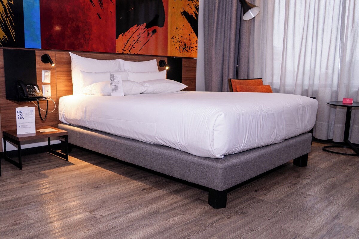 A modern hotel room with a large bed, a bedside telephone, a nightstand, a lamp, and a small table with a pink object.