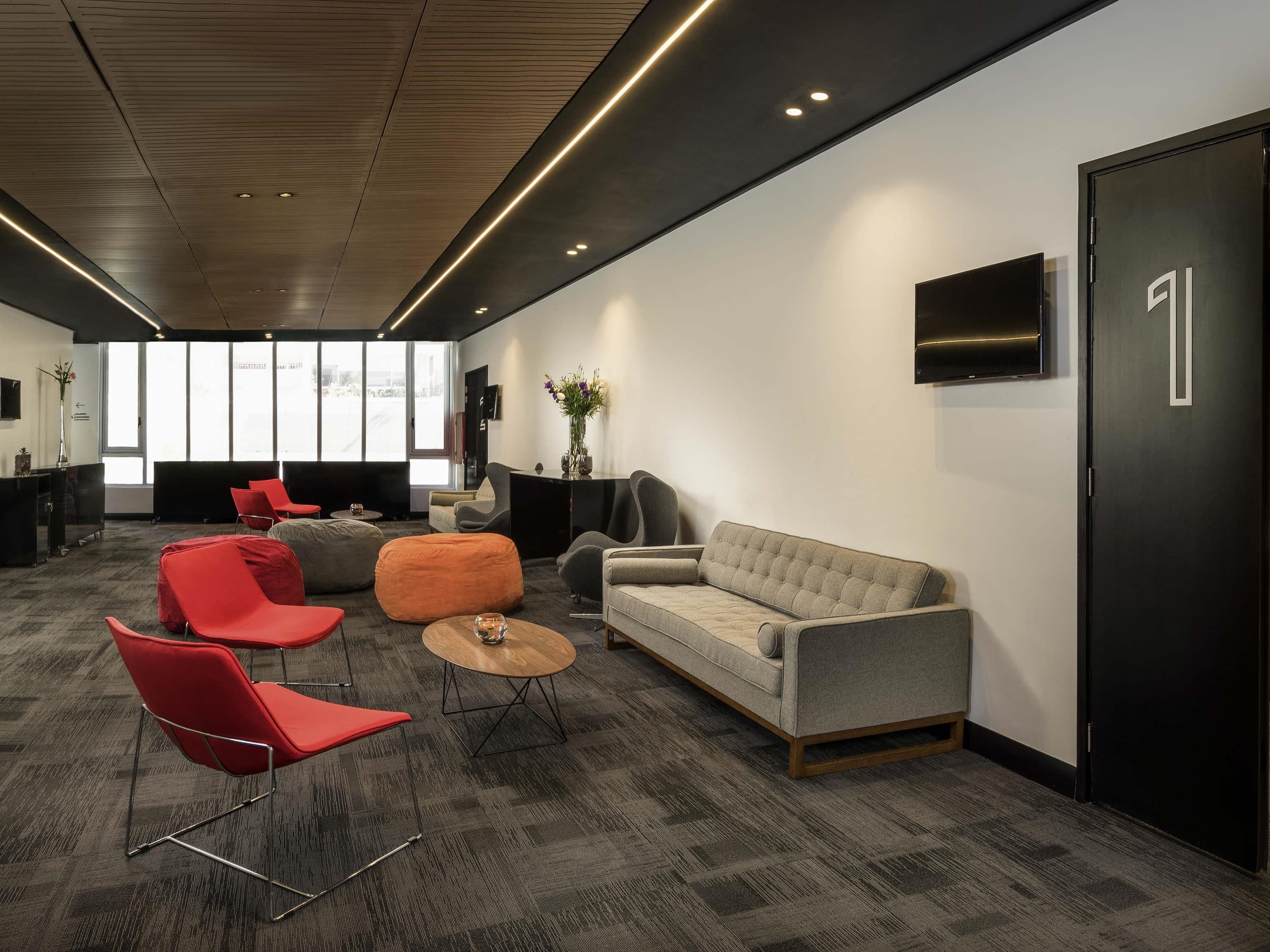 Un espacio de oficina moderno con un sofá gris, sillas rojas, una mesa de centro de madera y una puerta negra.
