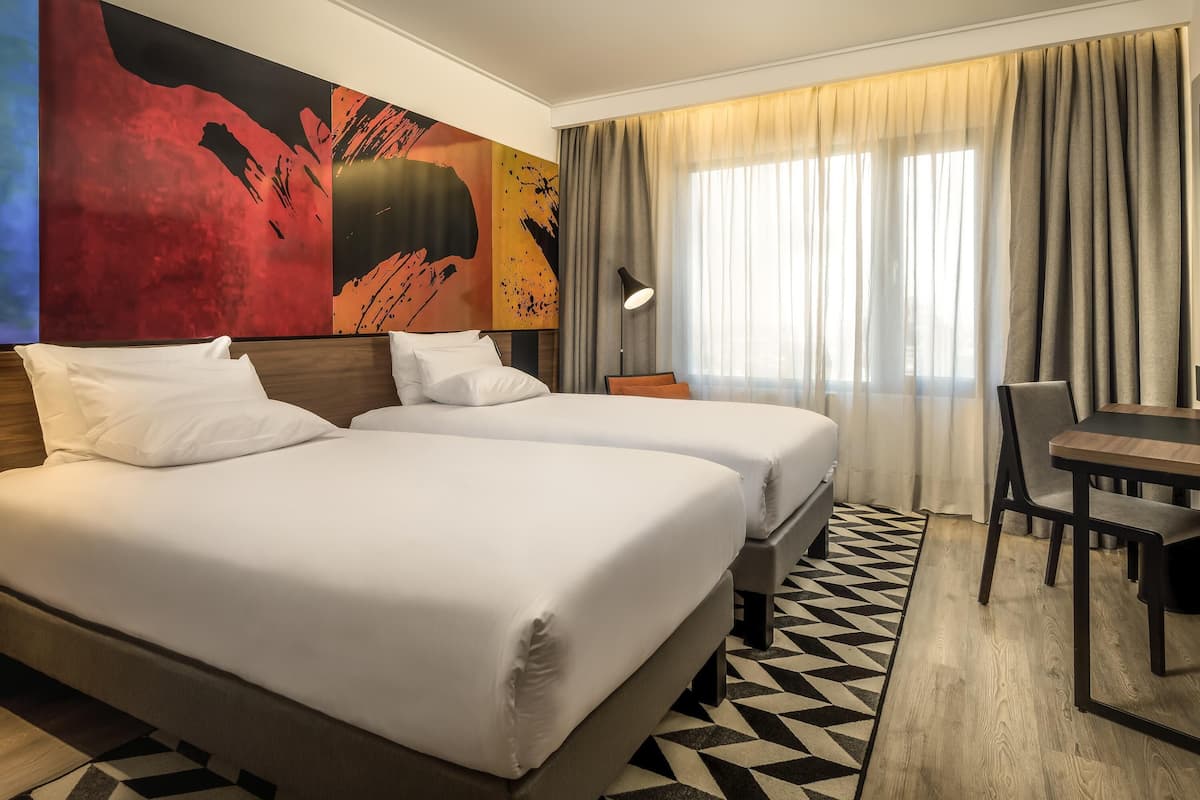Une chambre d’hôtel avec deux lits, un bureau, une lampe et un grand tableau abstrait accroché au mur.