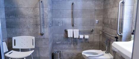 A bathroom with a wheelchair-accessible chair, grab bars, a toilet, and a trash can.