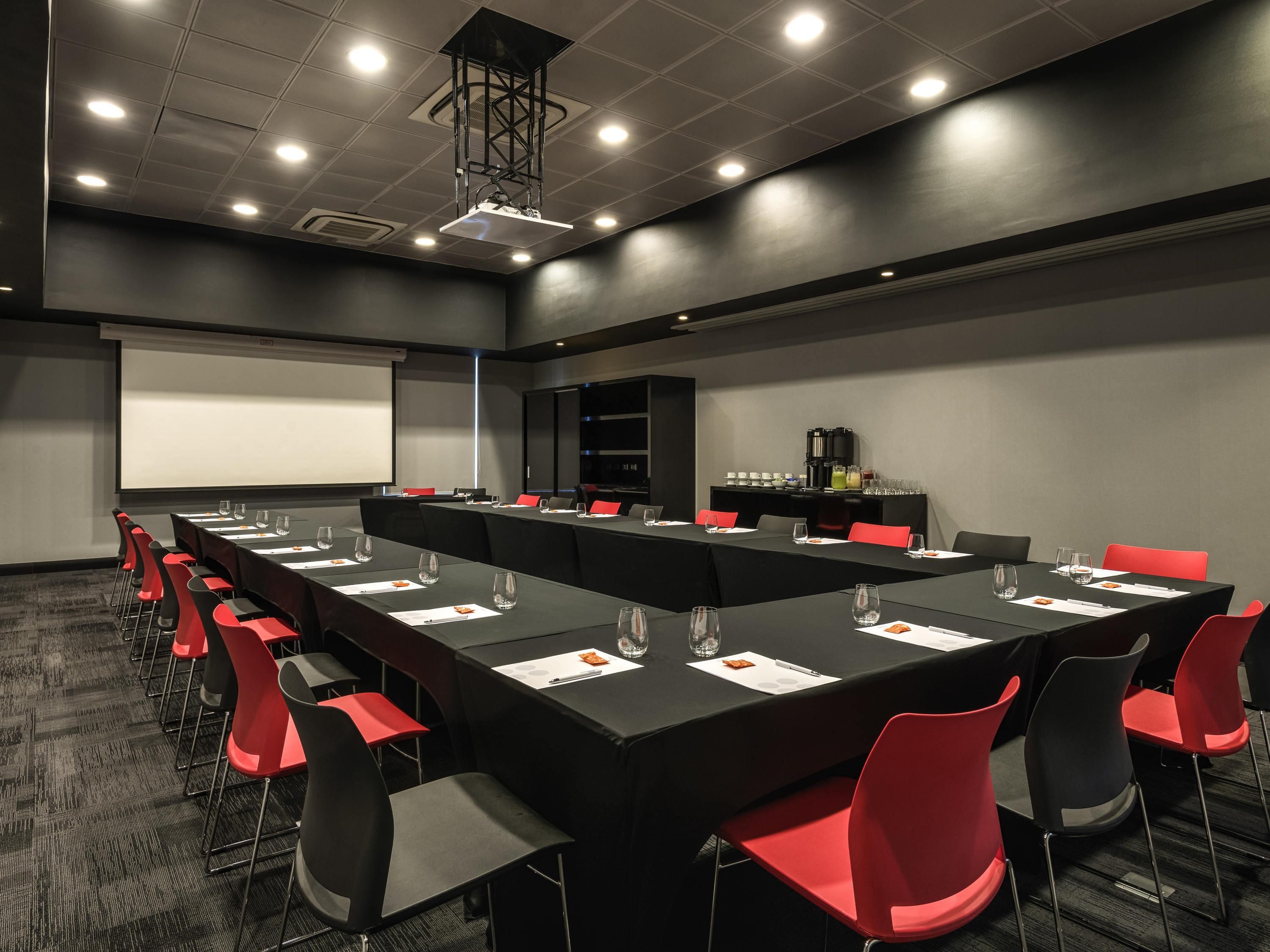 A conference room with a long table, red and black chairs, a projector screen, and a coffee station.