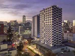 Un hotel moderno con fachada a cuadros, rodeado de edificios urbanos y una calle concurrida al anochecer.