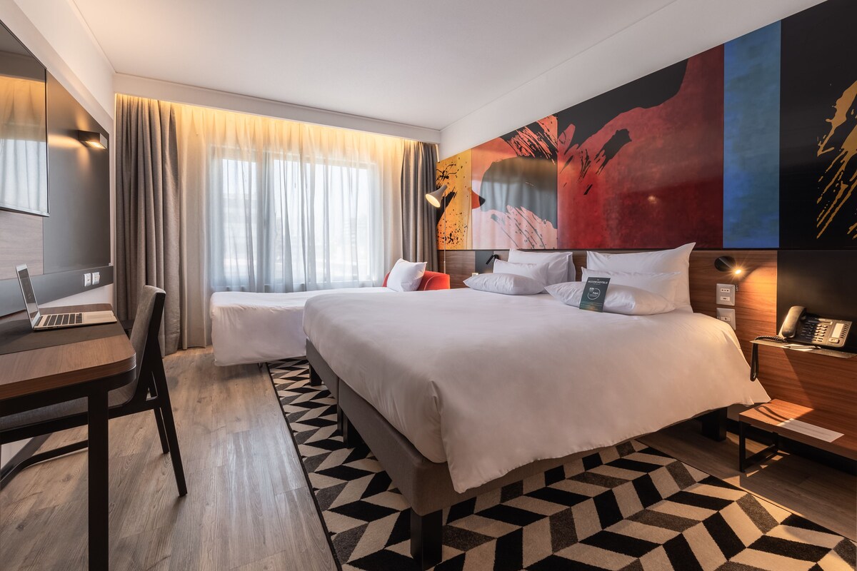 A hotel room with a large bed, a desk with a laptop, a chair, and a bedside table with a phone.