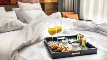 Una cama bien tendida con sábanas blancas, una bandeja con cruasanes, vasos de jugo y tazas, y un colorido cuadro abstracto al fondo.