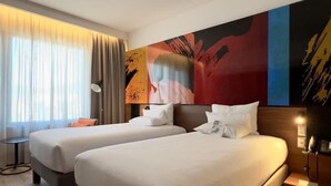 Quarto de hotel com duas camas, uma janela grande com cortinas, um quadro moderno na parede e uma mesa de cabeceira com abajur.
