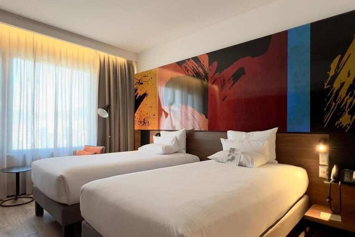 Une chambre d’hôtel avec deux lits, une grande fenêtre avec des rideaux, un tableau moderne accroché au mur et une table de chevet avec une lampe.