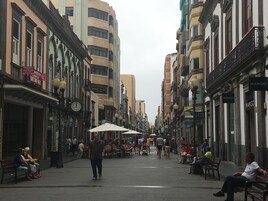 Calle flanqueada por edificios, algunos con letreros y anuncios. Hay bancos y gente caminando o sentada.
