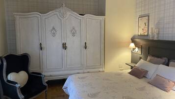Un dormitorio con un armario blanco grande, un sillón negro con un cojín con forma de corazón, una cama con ropa de cama floral y una mesita de noche con una lámpara.