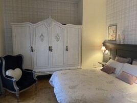 A bedroom with a large white wardrobe, a black armchair with a heart-shaped cushion, a bed with floral bedding, and a nightstand with a lamp.