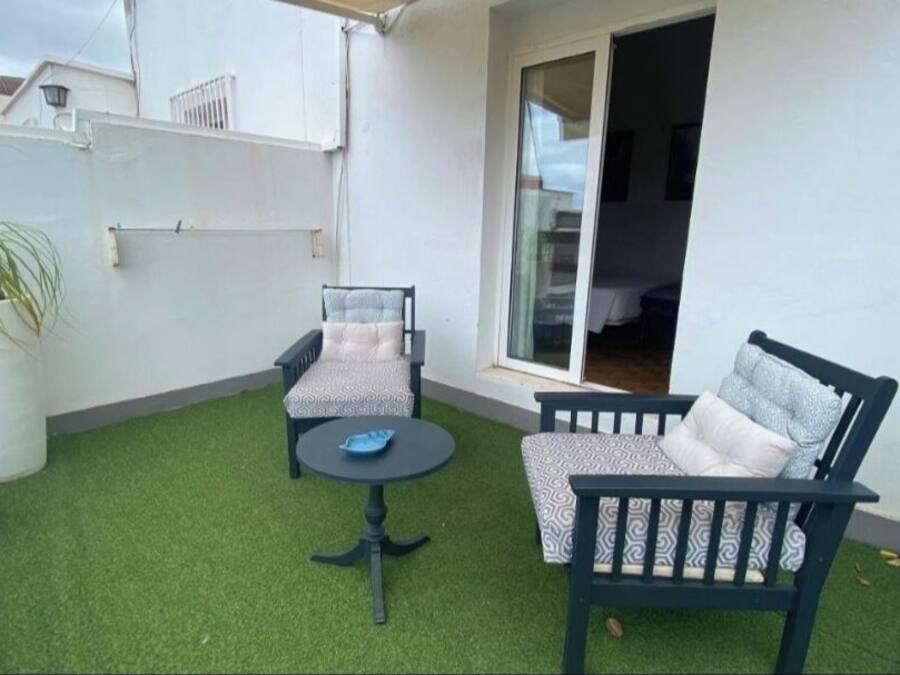 Een klein terras buiten met twee stoelen en een tafel, omringd door kunstgras en potplanten.