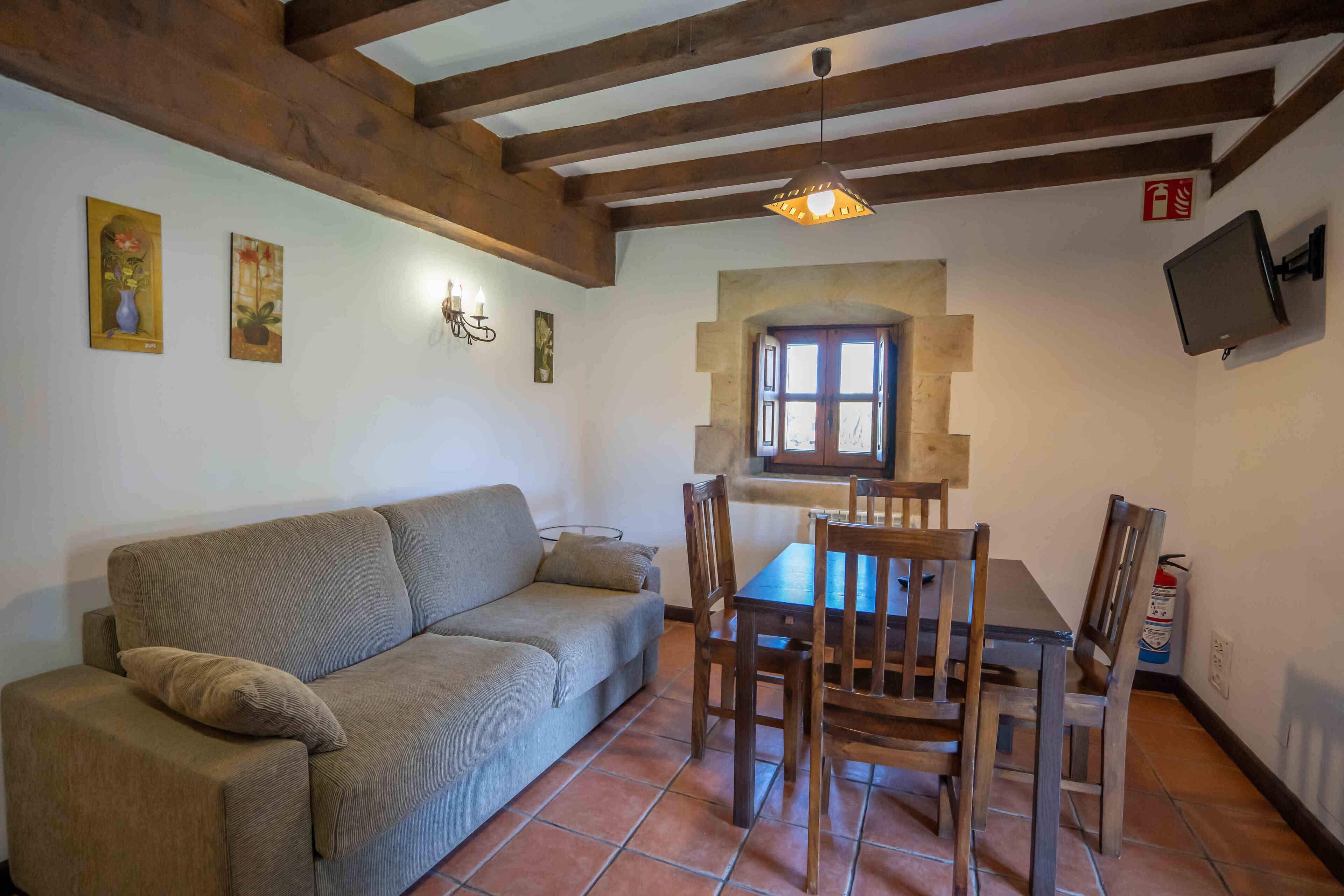 Un espacio acogedor con sofá, mesa de comedor de madera y sillas. Hay luces fijadas en la pared y un televisor.
