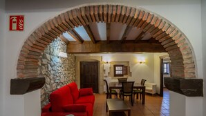 Un intérieur chaleureux avec un canapé rouge, une table à manger en bois et des chaises. On peut apercevoir un mur en pierre et une arche en briques.