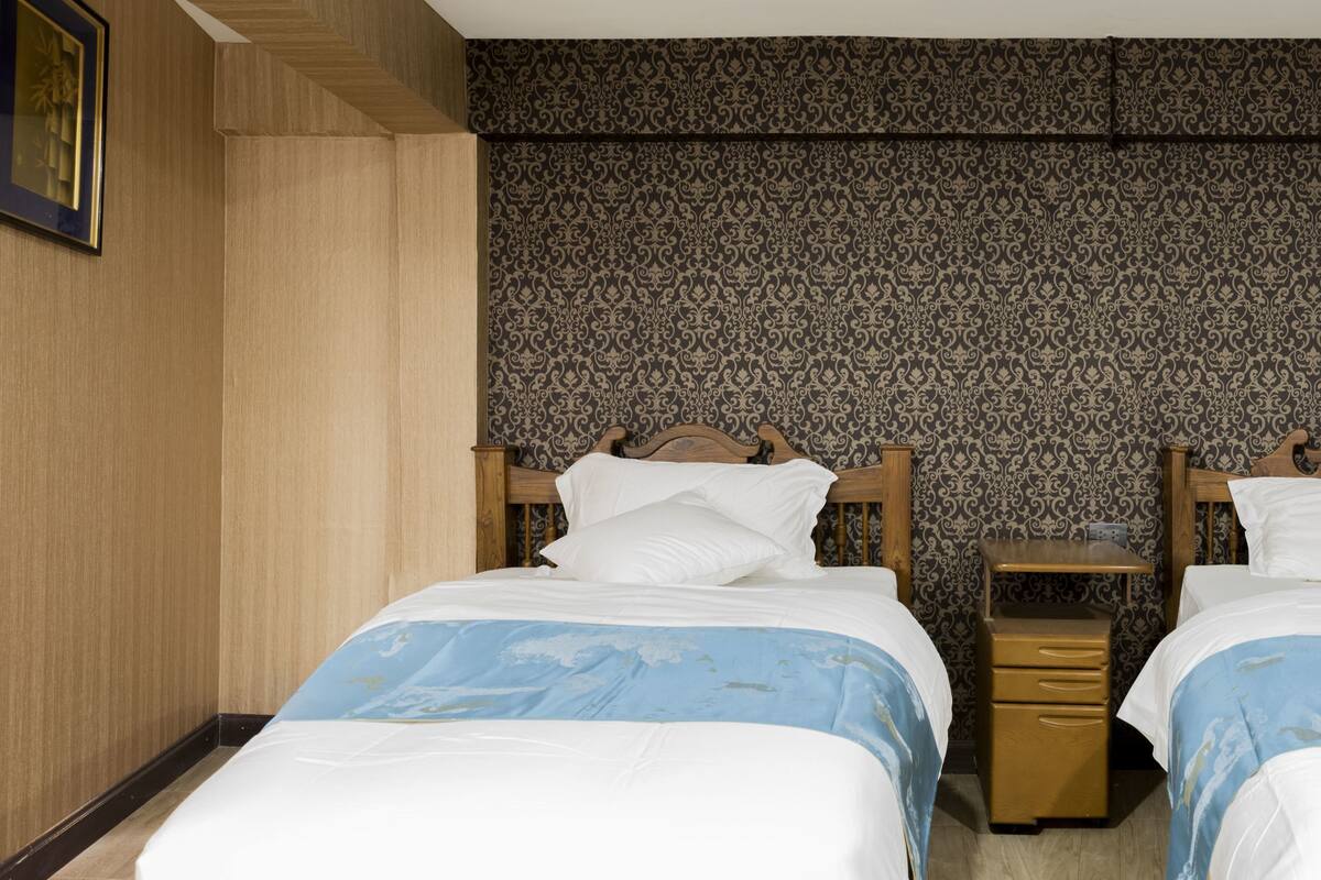 Een kamer met twee bedden, een klein houten nachtkastje, behang met patroon en een raam met gordijnen.