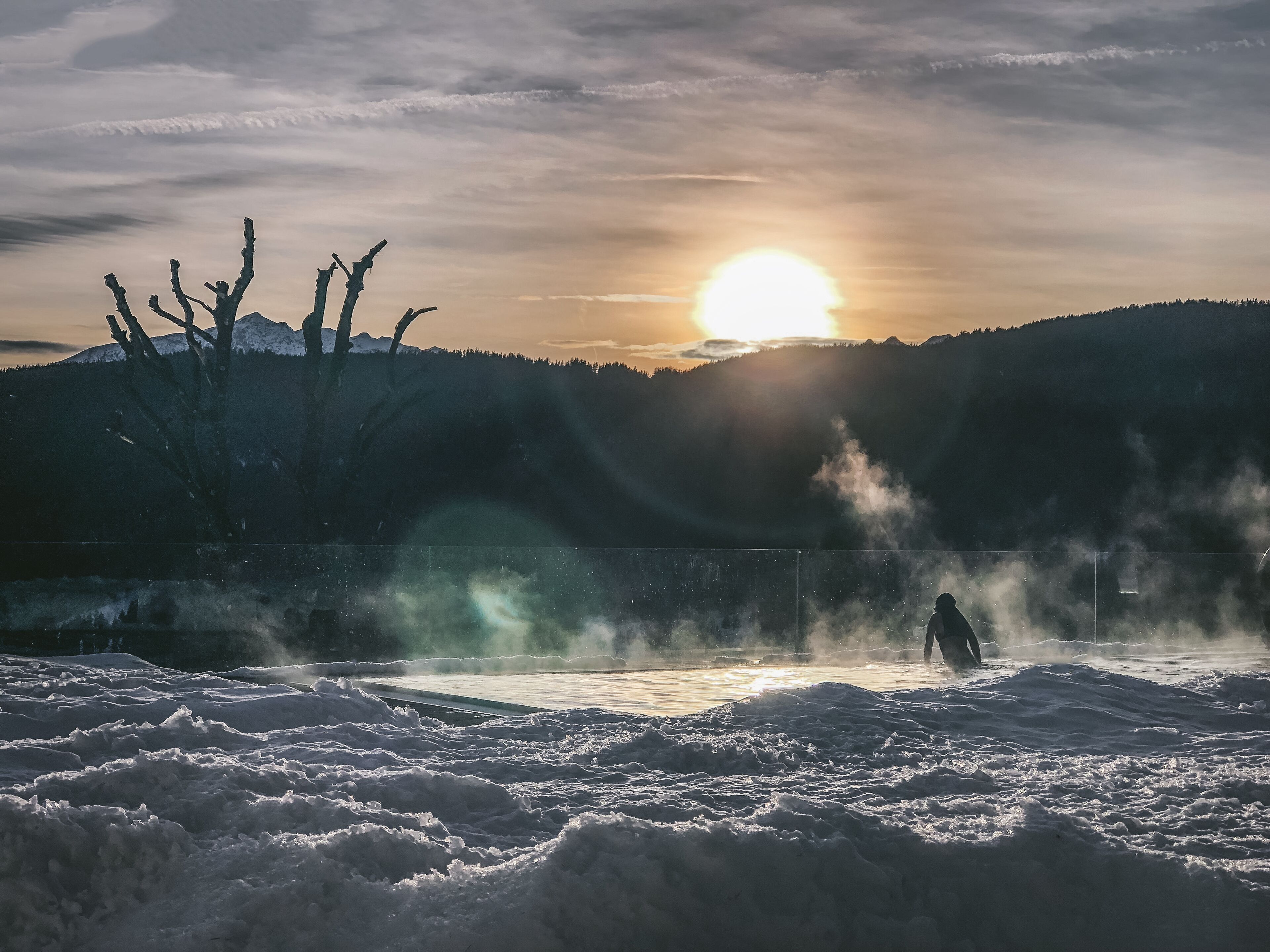 Un paysage enneigé avec une personne debout près d’une clôture, des montagnes en arrière-plan et le soleil se couchant ou se levant.