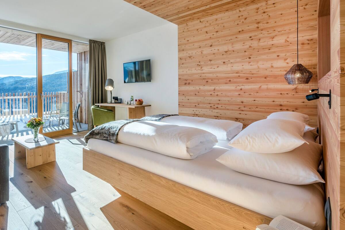 Une chambre d’hôtel moderne avec un intérieur en bois, un canapé-lit, un bureau avec une télévision et un balcon offrant une vue sur la montagne.