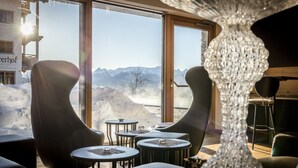 Een modern interieur met uitzicht op besneeuwde bergen door grote ramen. Er zijn gebogen zithoeken en kleine tafels met placemats.