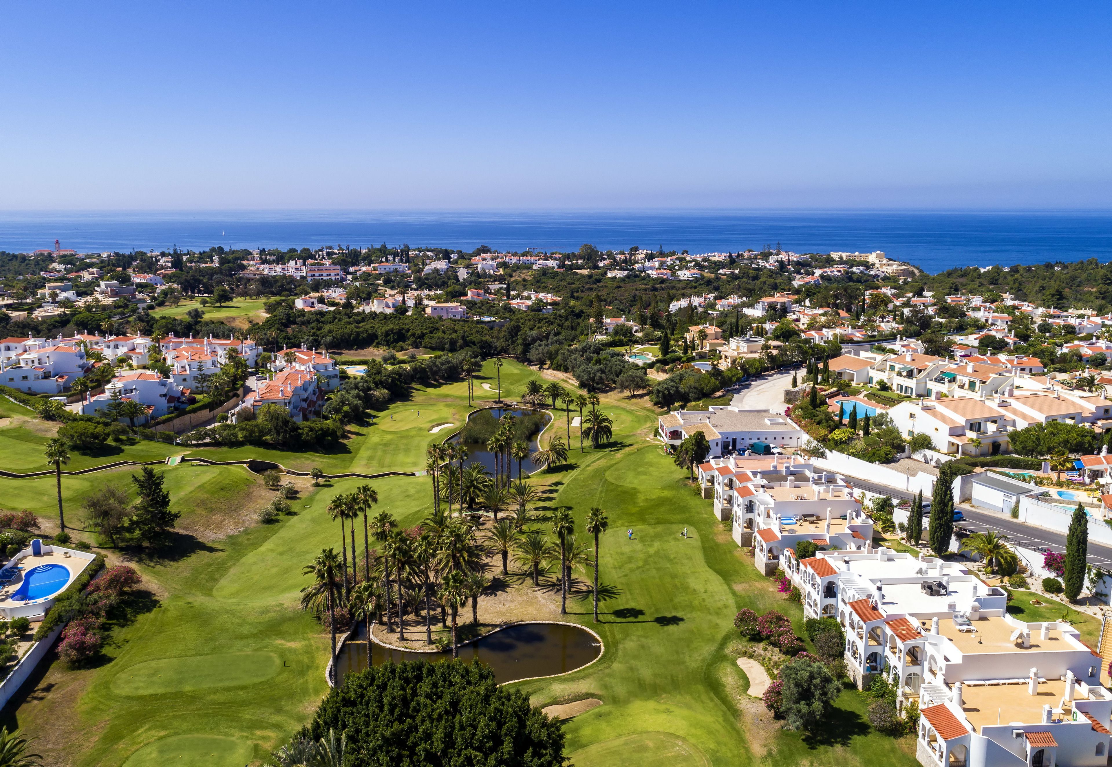 An aerial view of a coastal town with a golf course, residential buildings, and a clear view of the sea.