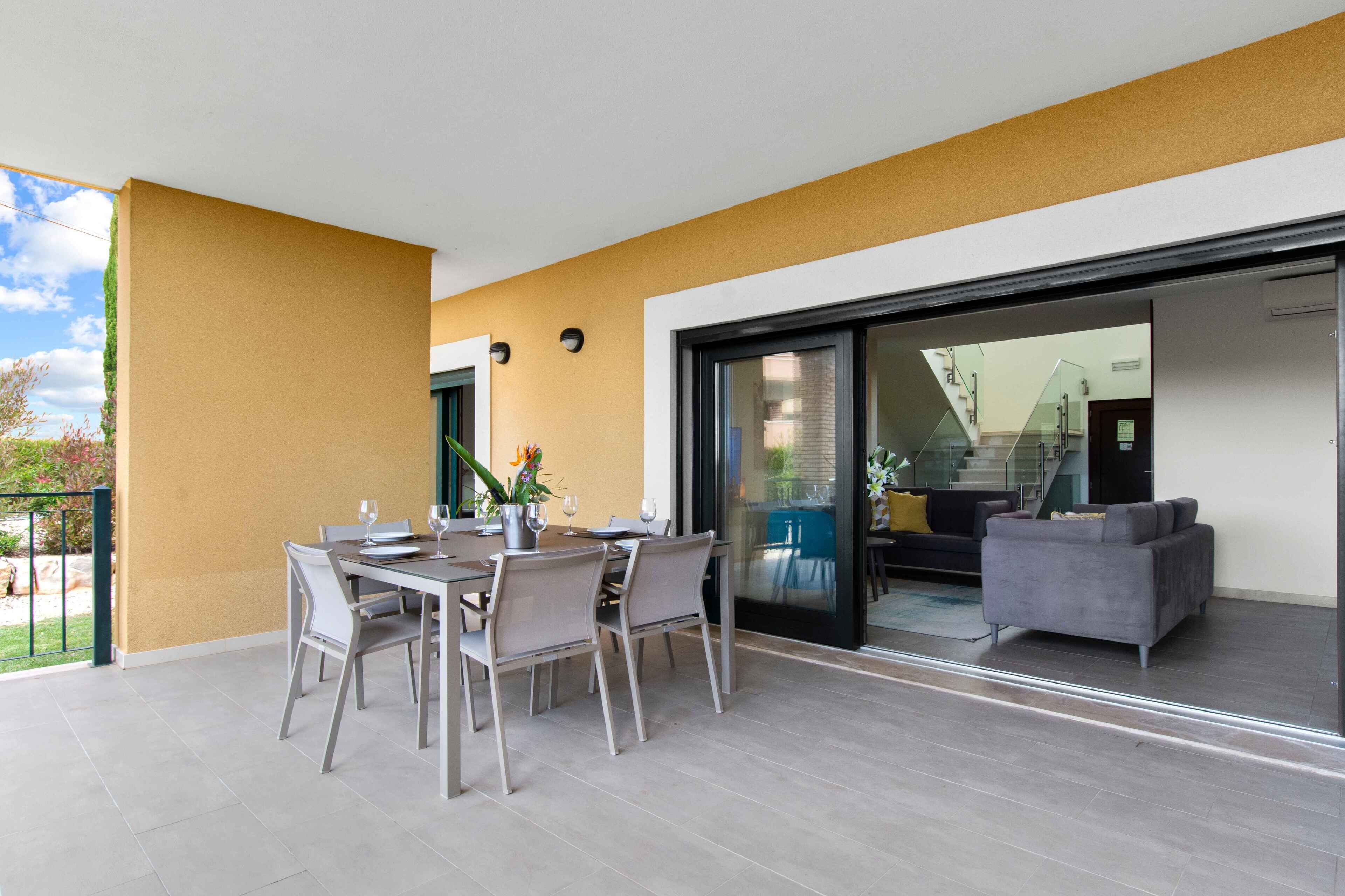 A modern outdoor dining area with a table and chairs, a sliding glass door leading to a living space, and a yellow wall.