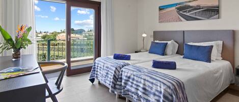 A hotel room with two beds, a dining table, a potted plant, and a balcony with a view.