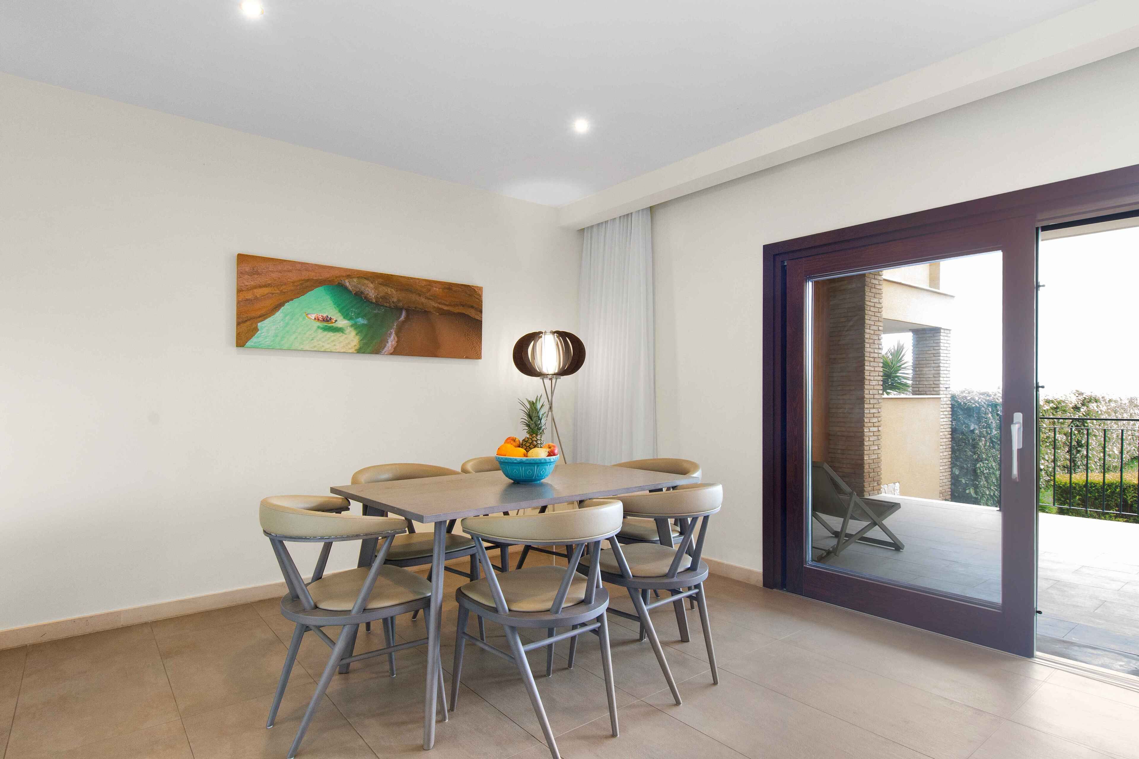 A dining area with a round table and six chairs, a painting on the wall, and a view through a glass door to an outdoor balcony.