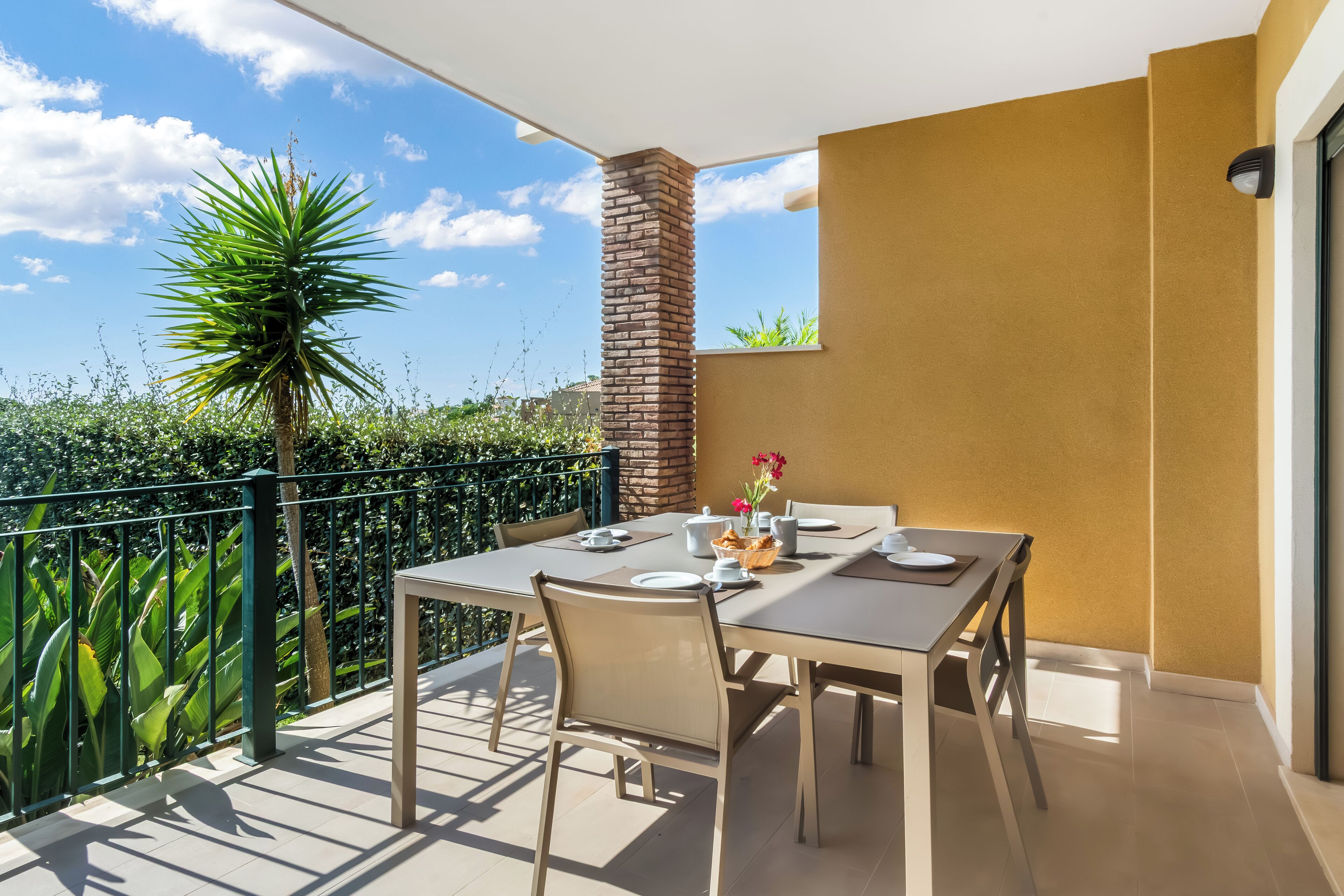Een balkon met een tafel gedekt voor twee personen, een bloempot en een uitzicht op de lucht en groen.