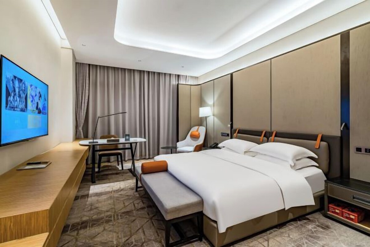 A modern hotel room with a large bed, a desk, a chair, a TV, and a large window with curtains.