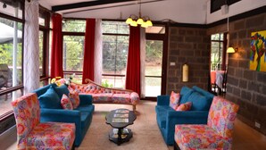 A cozy living room with blue sofas and colorful patterned chairs, a coffee table, and large windows with red curtains. - Kifaru House - Karen (Nairobi)