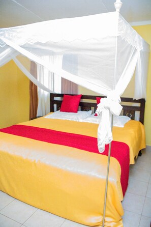 Een tweepersoonsbed met hemelbed, beddengoed rood en geel, en een witte gordijn.