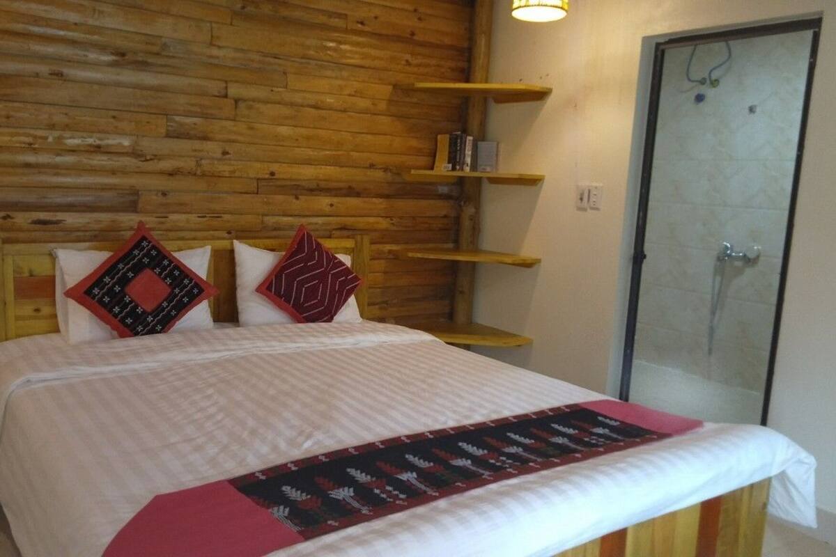 Une chambre à coucher avec une tête de lit en bois, un lit avec une literie à motifs, une table de chevet, une commode, une cabine de douche en verre et une étagère avec des livres.