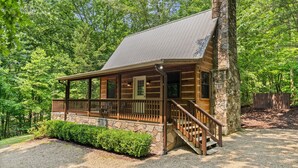 A log cabin with a stone chimney, wooden porch, and a gravel driveway.