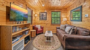 A cozy cabin interior with a wooden ceiling, a large flat-screen TV, a bookshelf, a red chair, a brown leather couch, and a round rug.