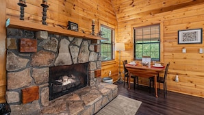 A log cabin interior with a stone fireplace, wooden walls, a dining area with a table and chairs, and a window with blinds.