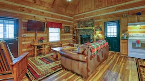 A log cabin interior with a leather couch, wooden walls, a stone fireplace, a television, and a kitchen area. - Bear Pause - 2 Bedrooms, 2 Baths, Sleeps 6 Cabin by RedAwning (Sevierville)