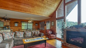 Ein gemütliches Blockhaus-Interieur mit einem Wohnzimmer, das eine Ledersofa, einen Couchtisch mit Zeitschriften, einen Kamin und große Fenster mit Blick ins Freie bietet.
