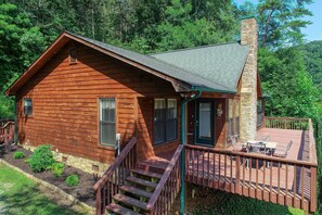 A wooden cabin with a stone chimney, a deck with outdoor seating, and a staircase leading to the entrance.
