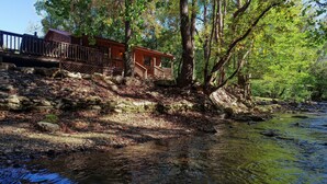 A cabin with a wooden deck and a stone wall, surrounded by trees and a stream. - Wandering Creek - 2 Bedrooms, 1 Baths, Sleeps 6 Cabin by RedAwning (Cosby)