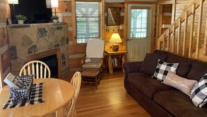A cozy cabin interior with a stone fireplace, wooden walls, a wooden dining table with checkered tablecloth, a brown sofa with checkered pillows, and a wooden staircase. - Sweet - 2 Bedrooms, 2 Baths, Sleeps 6 Cabin by RedAwning (Cosby)