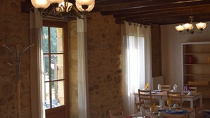 A dining area with wooden tables and chairs, a chandelier, and a view through a window with curtains. - Domaine Lacoste (Carsac-Aillac)
