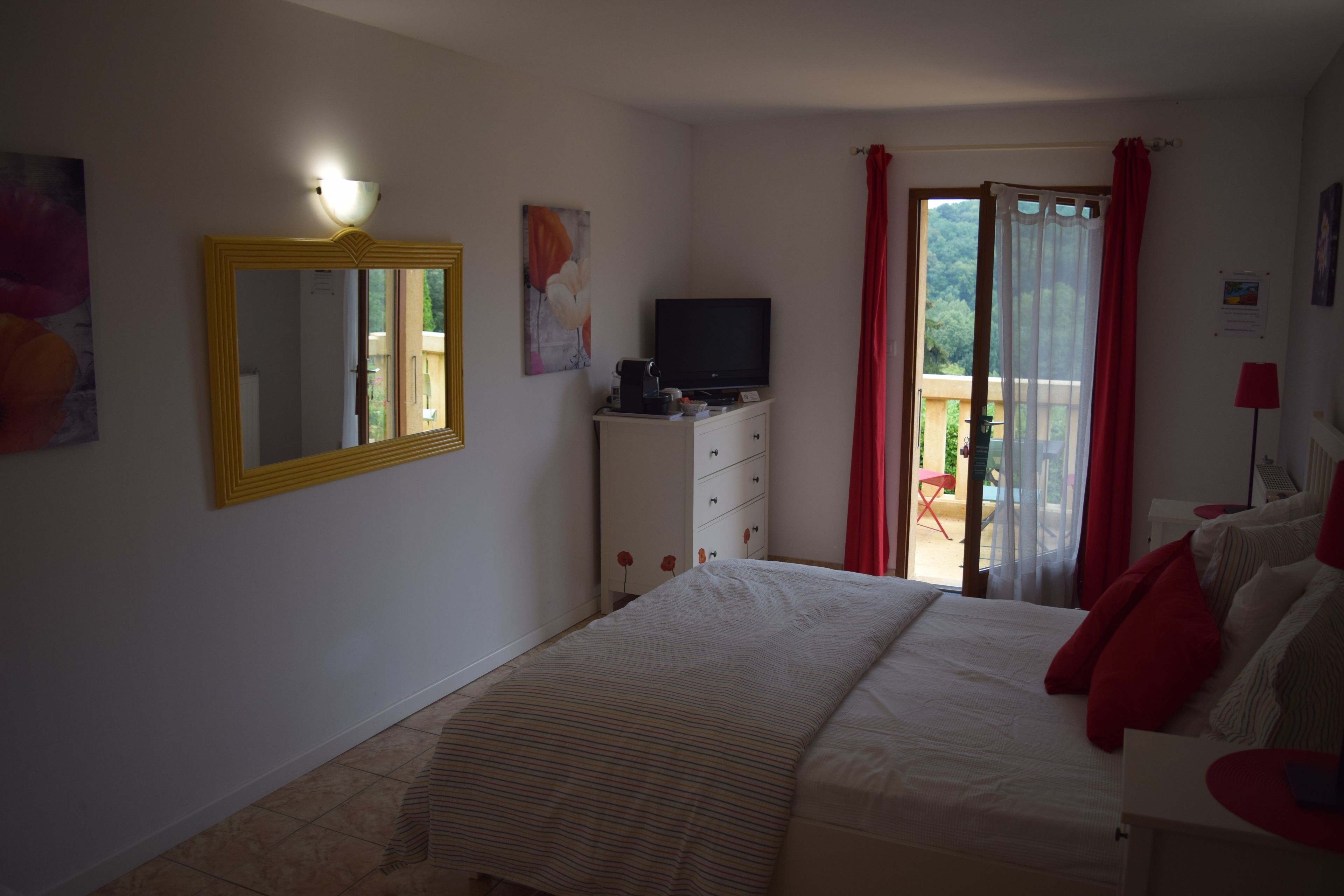 Un dormitorio con cama, cómoda, televisor y balcón con cortinas rojas.
