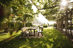 An outdoor patio area with tables and benches, surrounded by greenery and a view of a building in the background. - Hotel Bauhof (Castelbello-Ciardes)