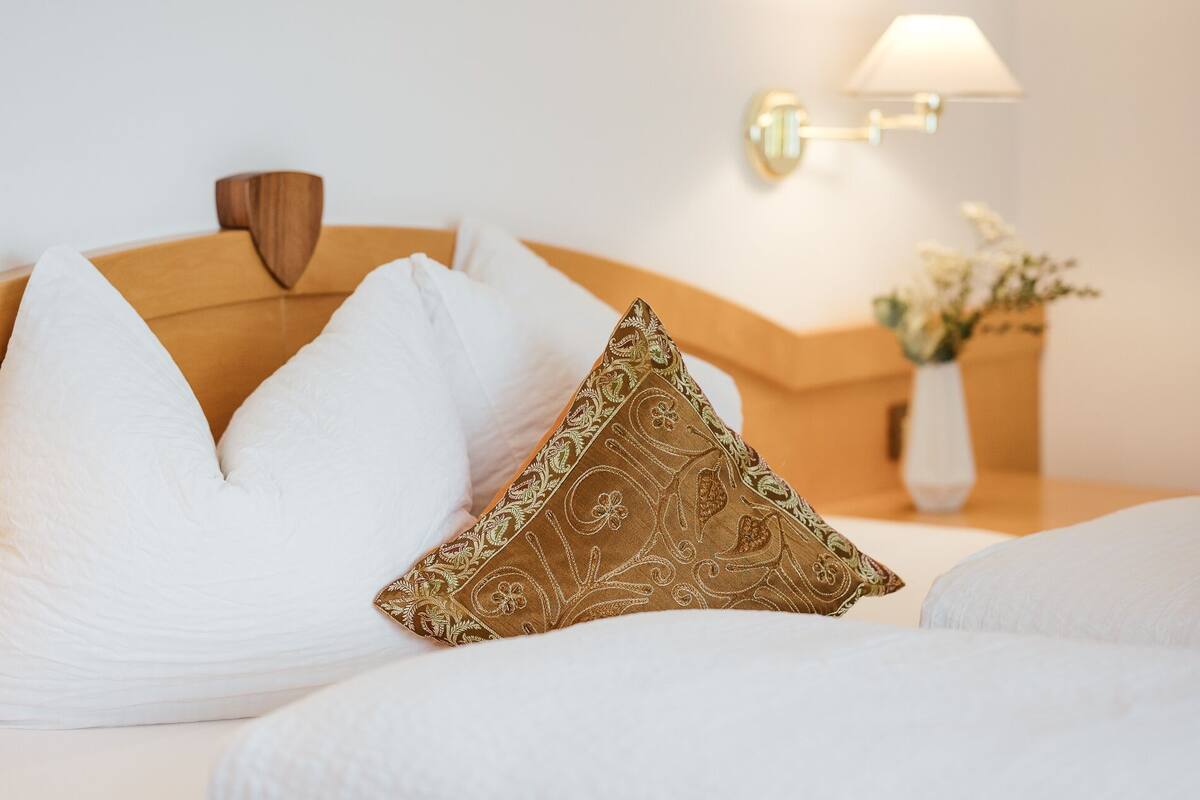 Un lit bien fait, avec des oreillers blancs et dorés, une tête de lit en bois et une table de chevet sur laquelle se trouvent une lampe et un vase de fleurs.