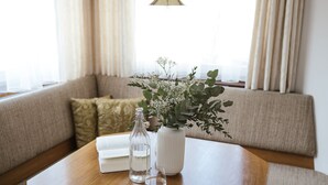 Et træbord med en hvid vase og to klare flasker, placeret på en sofa i et godt oplyst rum med et vindue og gardiner.