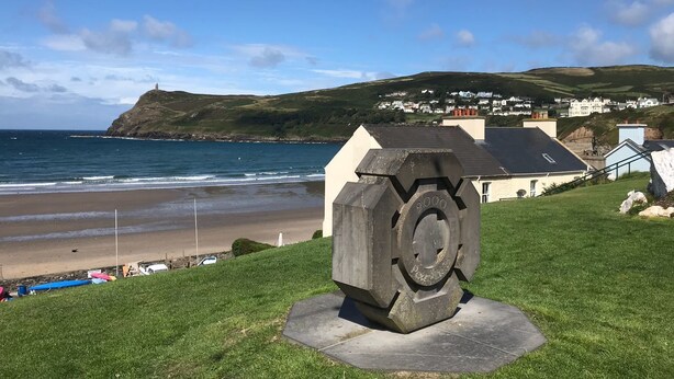 Un paysage côtier avec une zone herbeuse, une grande sculpture en pierre, une plage et une ville en arrière-plan.