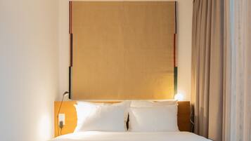 Une chambre d’hôtel avec une tête de lit en bois, un lit avec une literie blanche, une table de chevet avec une lampe et une fenêtre avec des rideaux.