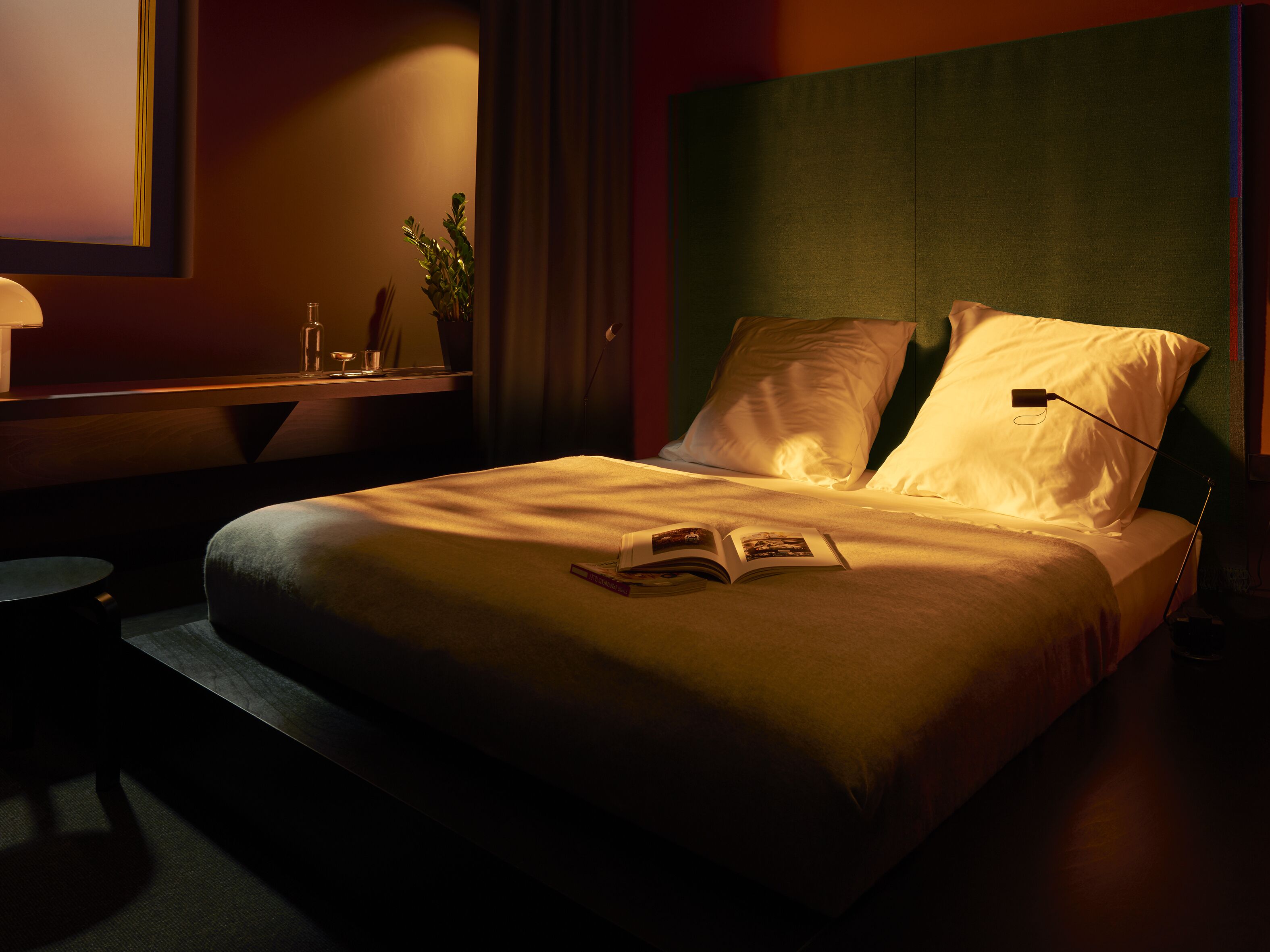 A neatly made bed with pillows and an open book, a bedside table with a lamp, and a small potted plant.