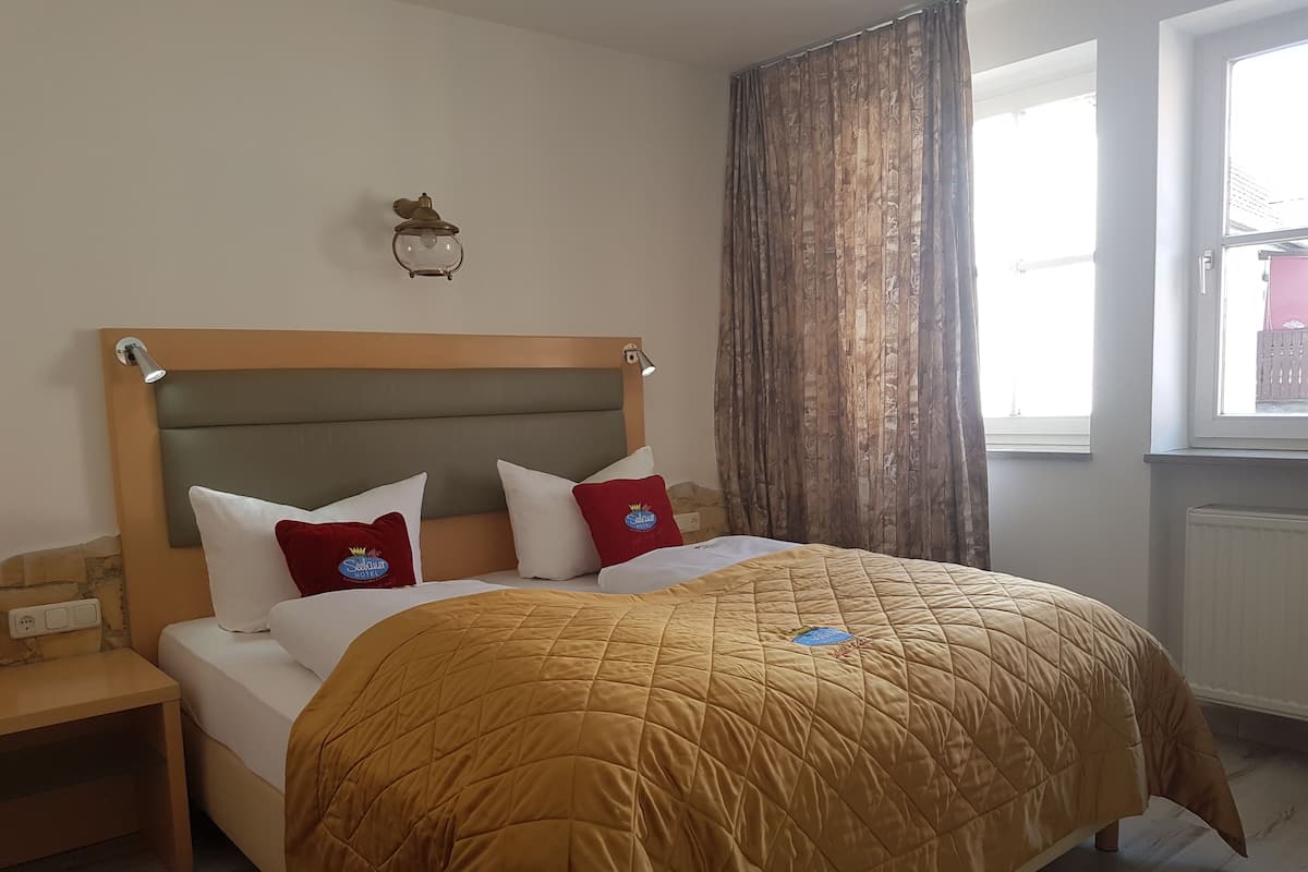 Una cama con cabecera, dos almohadas, una mesita de noche y una ventana con cortinas.